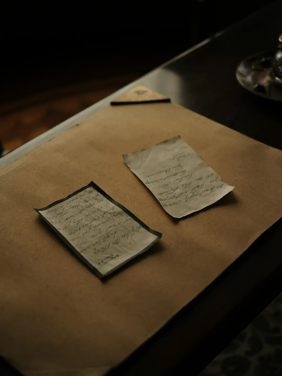 Duas folhas antigas manuscritas, com bordos gastos e vincos, pousadas sobre uma folha de papel pardo numa mesa escura